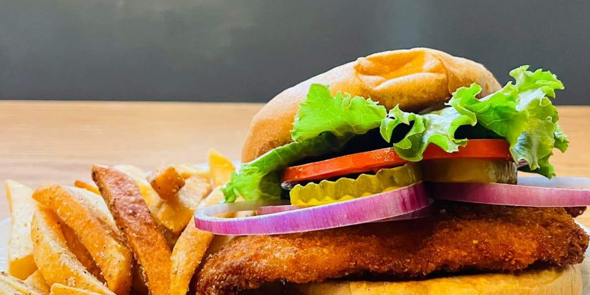 An appetizing image featuring a large fried sandwich and a pile of french fries on a white dish. The sandwich filling is breaded and golden, with fresh vegetables visible on top.