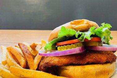 An appetizing image featuring a large fried sandwich and a pile of french fries on a white dish. The sandwich filling is breaded and golden, with fresh vegetables visible on top.