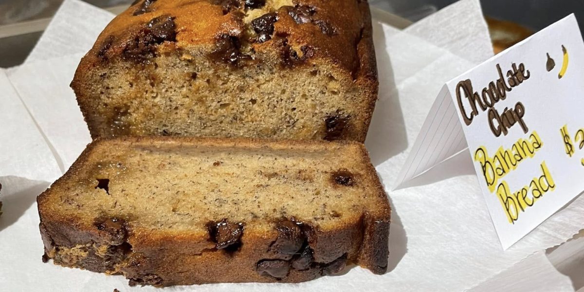 A loaf of chocolate chip banana bread, sliced open to show the inside, displayed on a tray with a small handwritten sign indicating it's "Chocolate Chip Banana Bread" and its price.