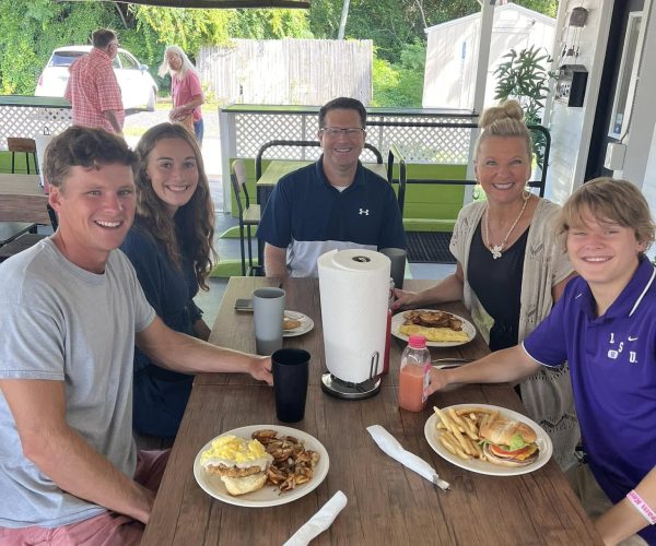 A family of five smiling and enjoying a meal together at a wooden table on the covered patio of the Oh Biscuit restaurant.