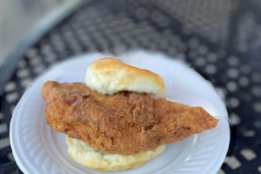 A close-up shot of a golden-brown, fried pork chop served inside a fluffy biscuit on a small white plate, resting on a black mesh outdoor table.