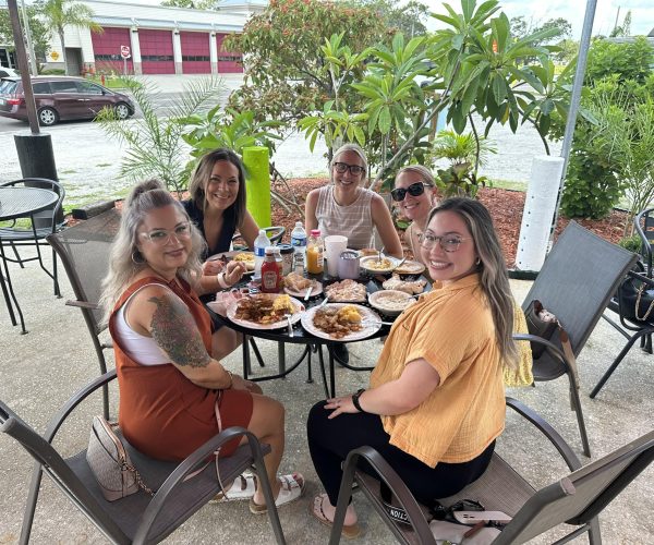 Five friends, all women, smiling and gathered around a round table filled with plates of food and drinks on the outdoor patio of the Oh Biscuit restaurant.