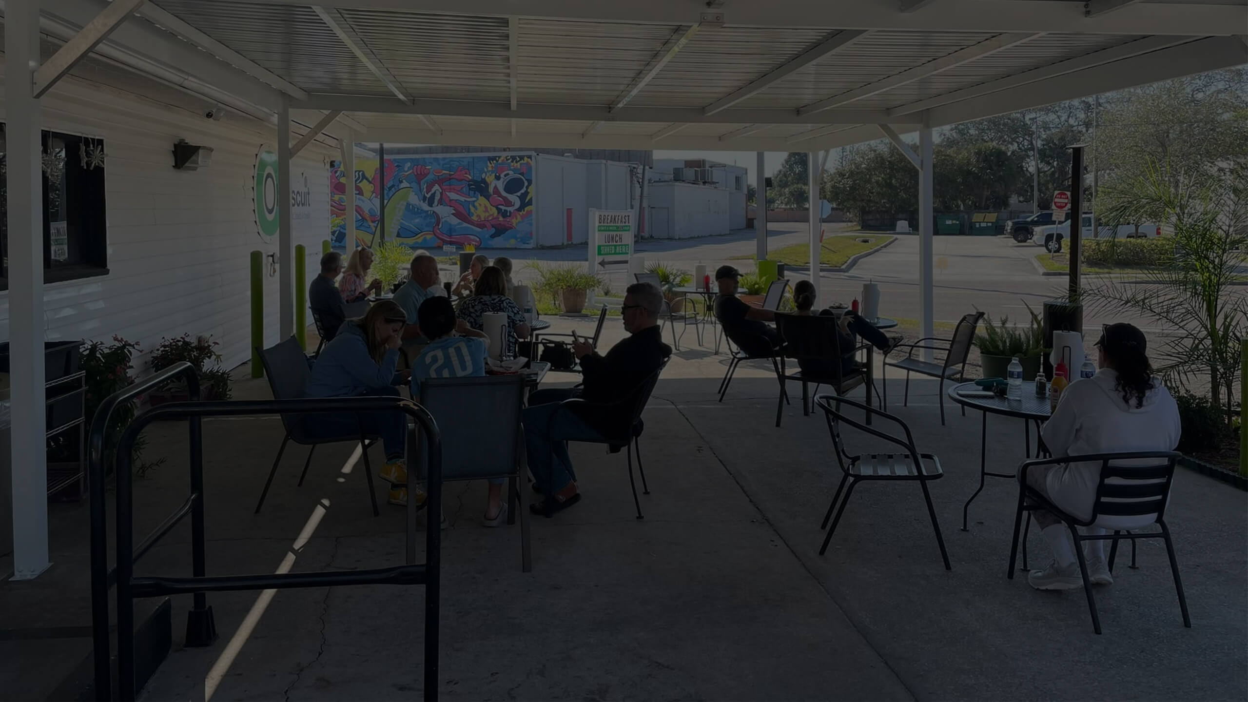 A wide shot of the covered outdoor patio at Oh Biscuit, with several customers seated at tables enjoying their meals on a sunny day. A colorful mural is visible on a building across the parking lot.