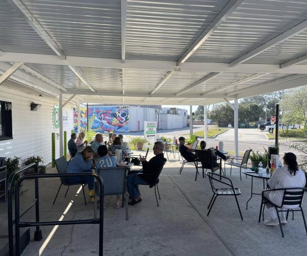 A wide shot of the covered outdoor patio at Oh Biscuit, with several customers seated at tables enjoying their meals on a sunny day. A colorful mural is visible on a building across the parking lot.