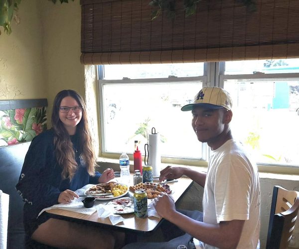 Two students, a young woman and a young man, sitting at a table by a window inside the Oh Biscuit restaurant, smiling at the camera while enjoying their food and canned drinks.