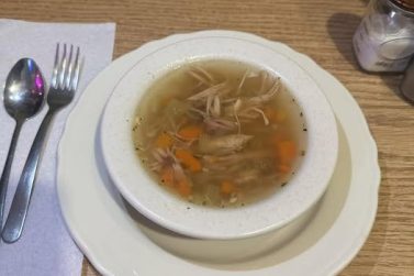 A bowl of hot chicken noodle soup with shredded chicken, carrots, and celery, served on a white plate with a spoon and fork on a paper napkin on a wooden table.