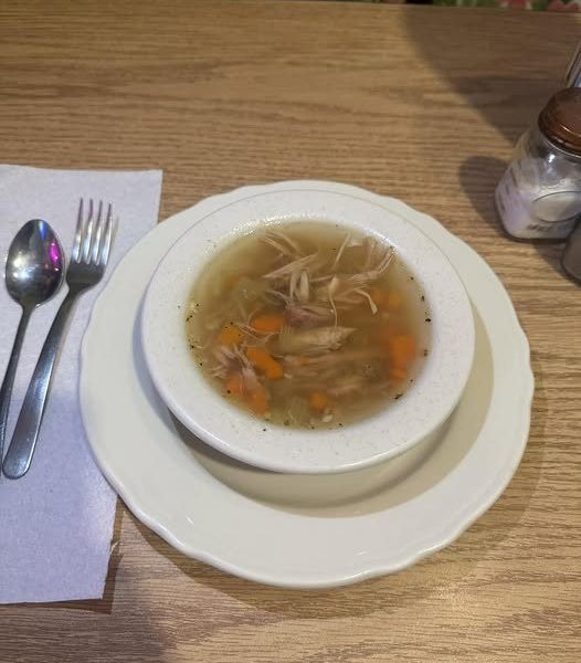 A bowl of hot chicken noodle soup with shredded chicken, carrots, and celery, served on a white plate with a spoon and fork on a paper napkin on a wooden table.