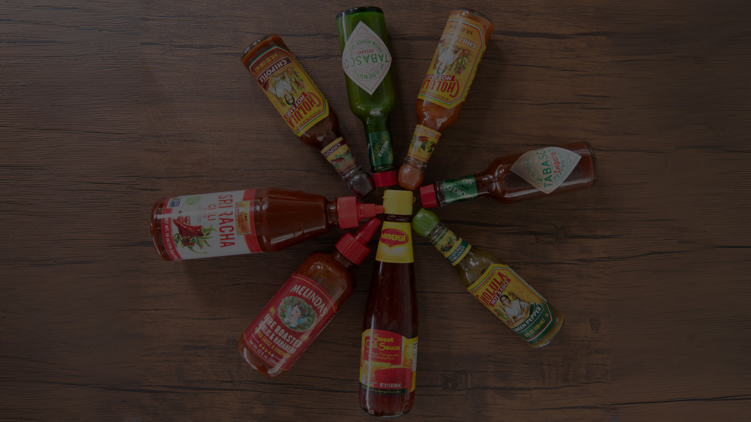 A top-down view of seven different hot sauce bottles, including Sriracha, Tabasco, Cholula, and Melinda's, arranged in a circular pattern on a wooden table.