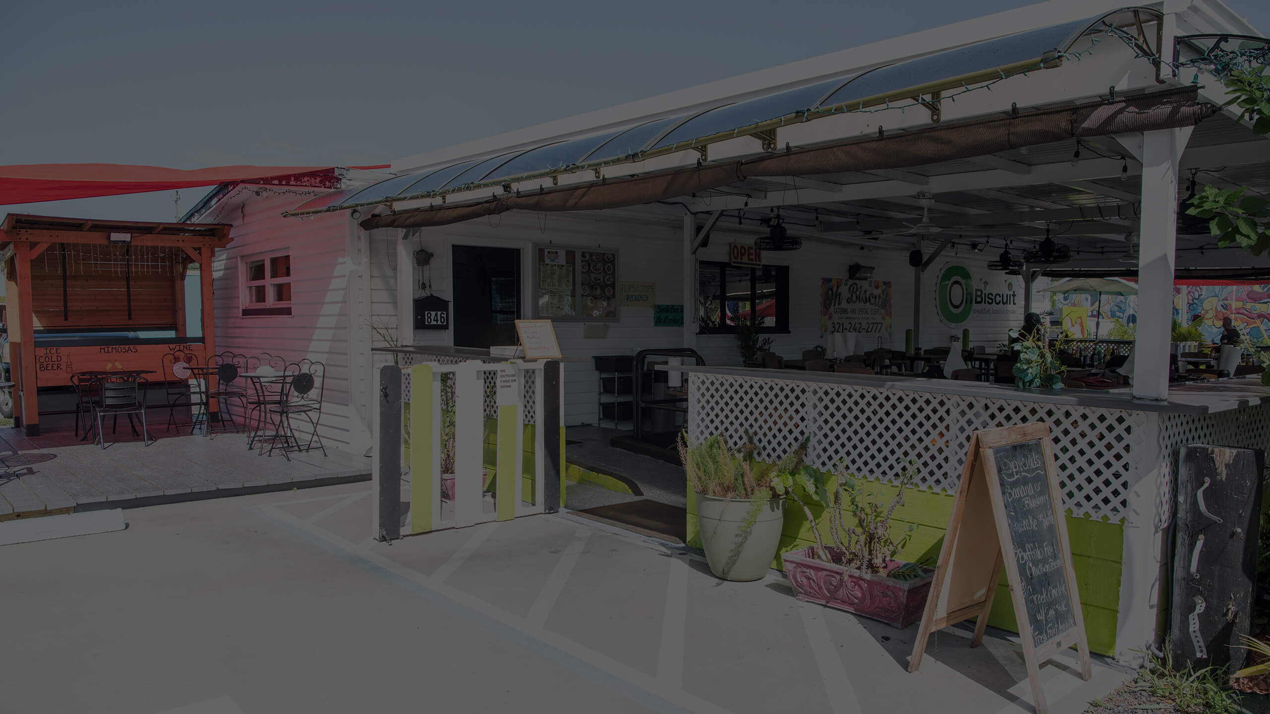 The white-painted exterior of the Oh Biscuit restaurant, showcasing its covered outdoor patio with tables and chairs, and the entrance with a wooden A-frame sign. The restaurant's logo is visible on the building.