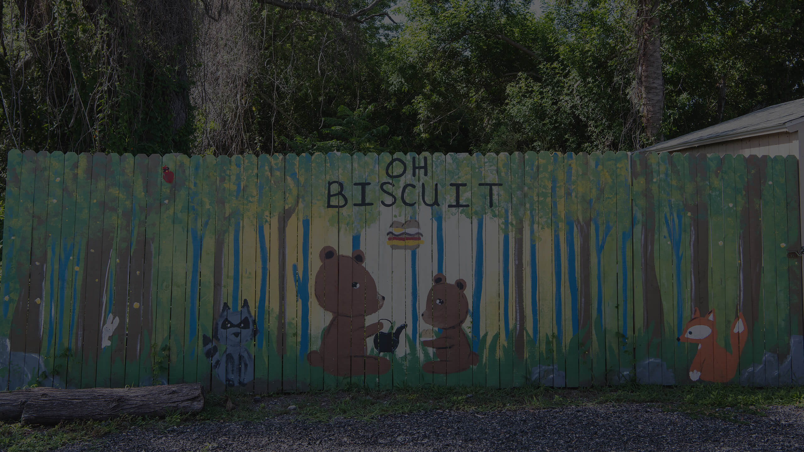 A colorful wooden fence outside the Oh Biscuit restaurant, painted with a whimsical mural of two bears sharing a cup of tea in a forest setting with a raccoon and a fox. The words "OH BISCUIT" are painted above the bears.