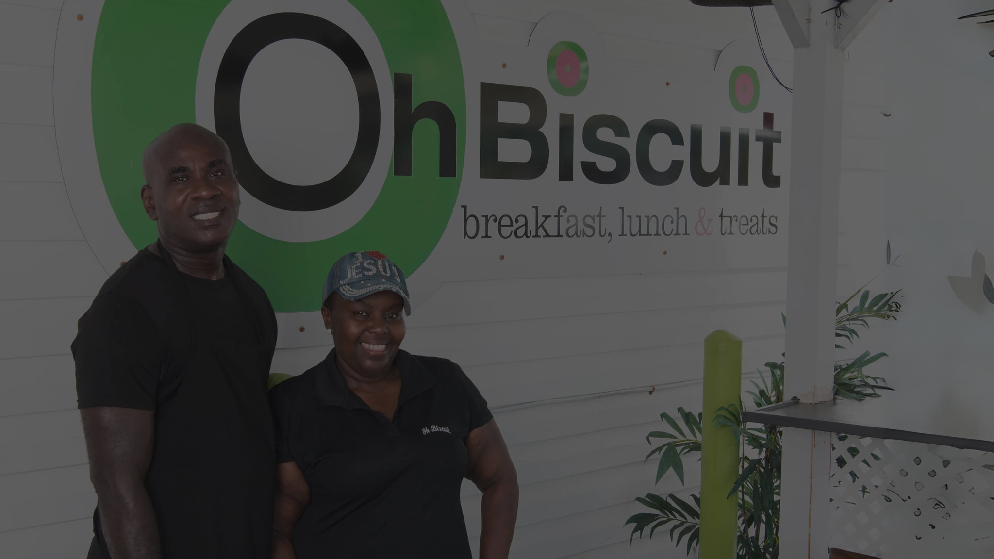 The two owners of Oh Biscuit, a man and a woman, standing side-by-side and smiling in front of the restaurant's main sign that reads "Oh Biscuit breakfast, lunch & treats."
