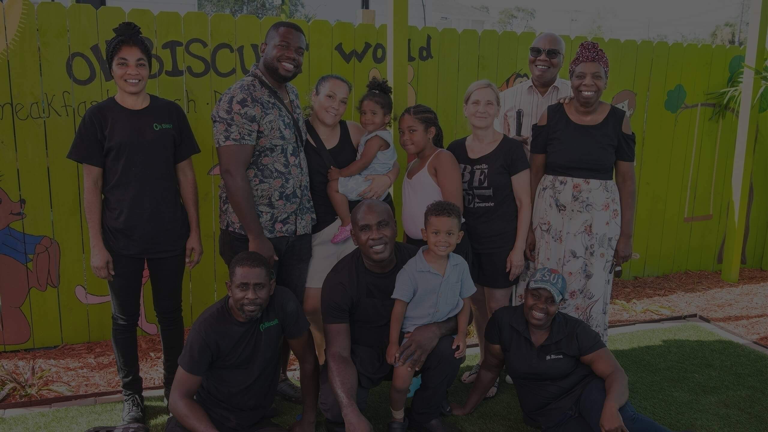 A group of people, including the owners, employees, and children, posing together for a group photo in the outdoor seating area of the Oh Biscuit restaurant in front of a green fence with a mural.